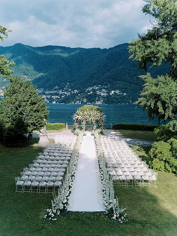 Outdoor ceremony setup with a floral arch and white aisle runner lined with flowers, facing a lake and mountain backdrop under cloudy sky