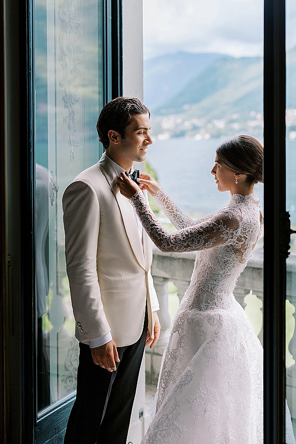 First look moment as bride fixing groom bow tie, her lace long sleeve dress and earrings framed by doorway to lake and mountains