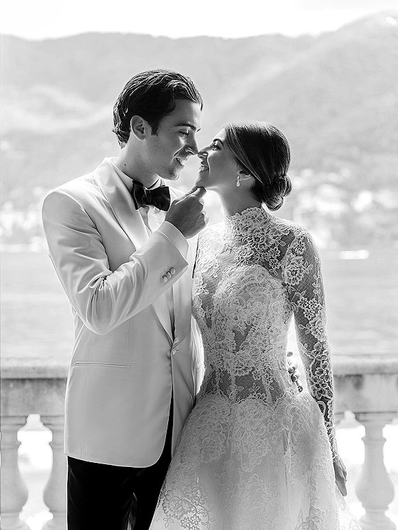 Couple portrait in black and white, groom touching bride’s chin as they lean forehead to forehead on a stone balcony by a lake and mountains