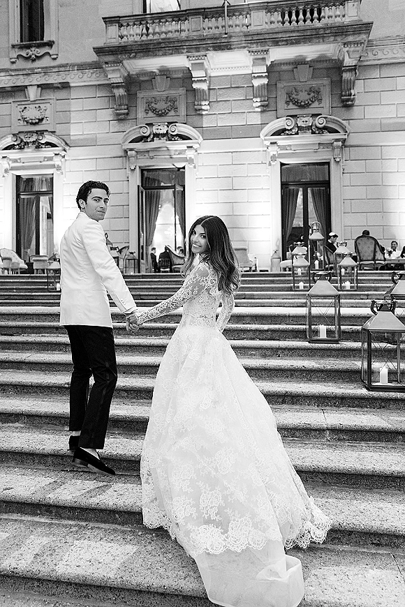 Couple portrait of bride and groom holding hands on stone steps, her long sleeve lace dress with train, lanterns glowing by grand facade