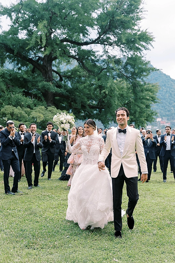 Wedding recessional as bride and groom walk hand in hand, bouquet raised, guests cheering on a lawn beneath a large tree with mountains behind