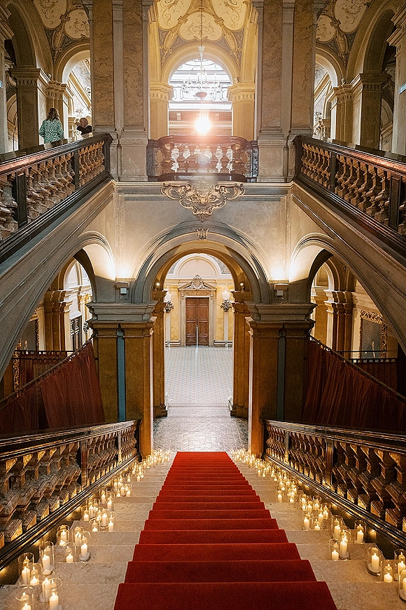 Wedding aisle decor with a red carpet runner lined by glass votive and pillar candles leading to a grand marble staircase under chandeliers