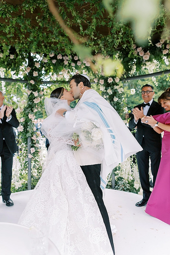 Wedding kiss as the couple kissing under chuppah, bride in lace veil holding white rose bouquet, groom in tallit, garden guests clapping behind