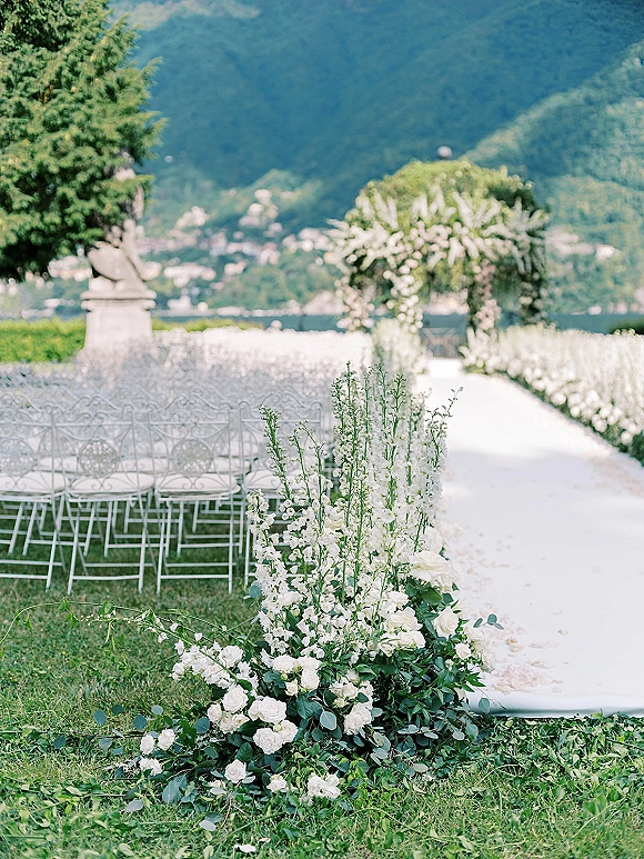 Ceremony aisle decor with an outdoor ceremony aisle lined by a white aisle runner, tall white florals and greenery, facing a floral arch by the lake and mountains