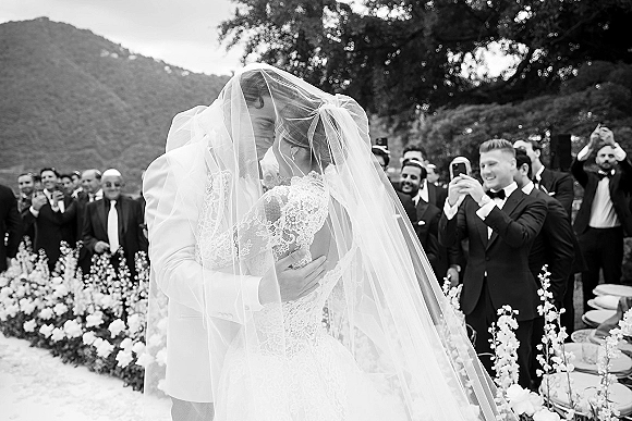 Wedding kiss under a veil over couple kiss, bride in lace dress and groom in white suit as guests cheer along a floral aisle outdoors