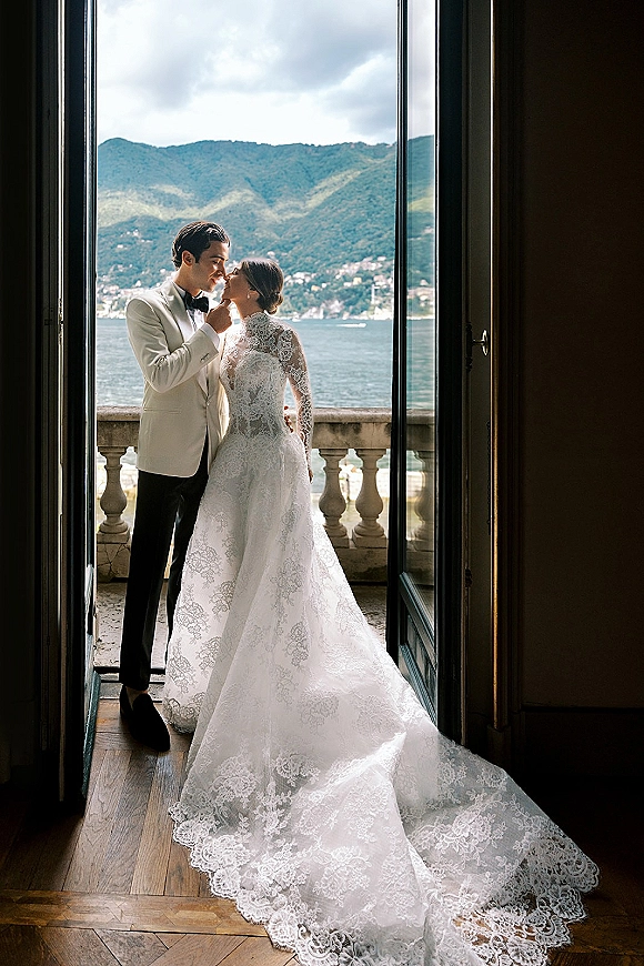 Couple portrait of bride and groom by window, forehead touching in doorway with lace gown and white tux, lake and mountains beyond