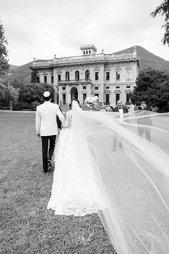 Wedding couple portrait of bride and groom walking away, bride’s long veil trailing as they cross a lawn by a historic facade, mountains beyond