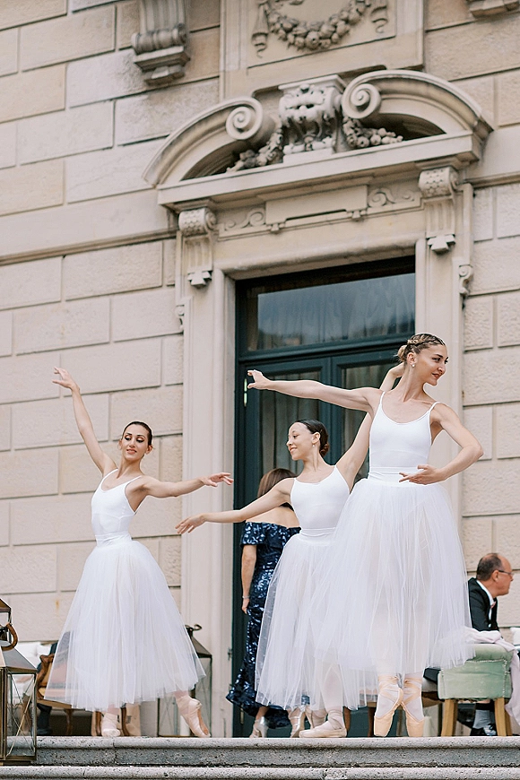 Wedding ballet performance with dancers in white tulle skirts and slippers on stone steps by a doorway, guests seated nearby outdoors