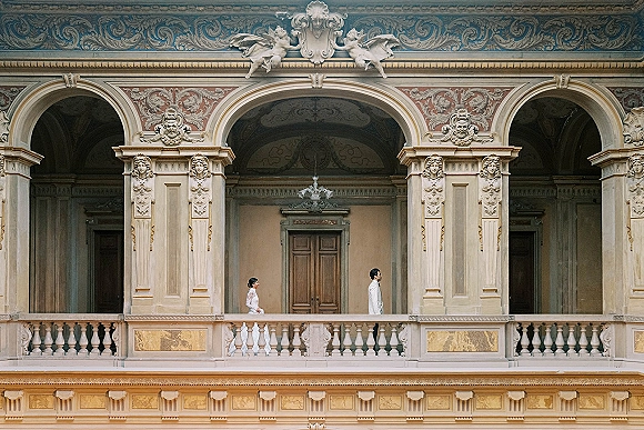 Couple portrait in an architectural wedding portrait style, bride in long-sleeve gown and groom in suit under an ornate arched colonnade balcony