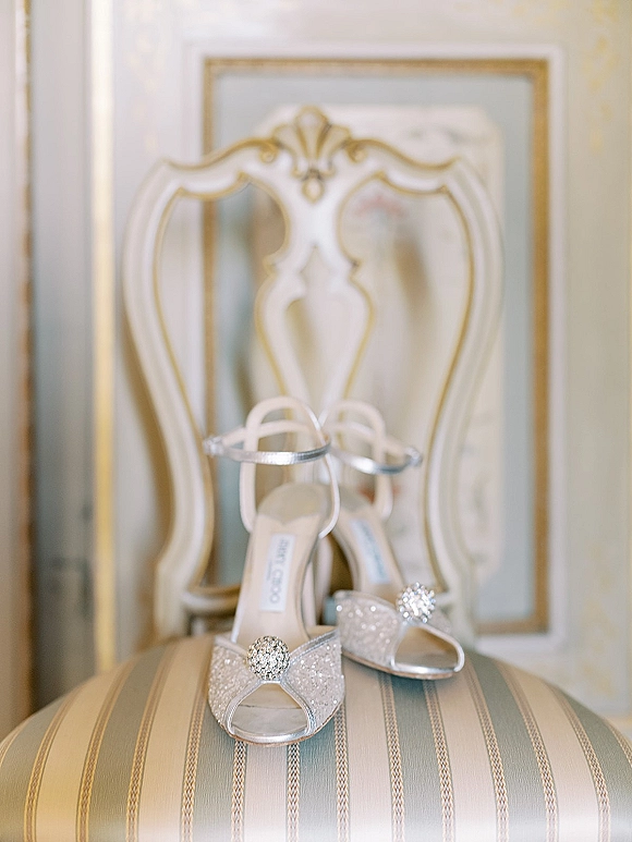 Wedding shoes with silver bridal shoes glitter fabric, rhinestone brooch and ankle straps resting on an ornate striped chair