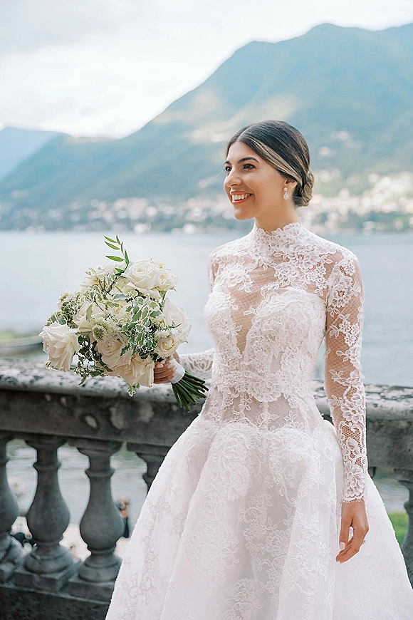 Bridal portrait of a bride holding bouquet of white roses in a high neck lace wedding dress by a stone balustrade with lake and mountains behind