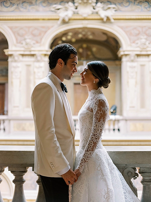 Couple portrait of bride and groom holding hands face to face, her lace long sleeve gown beside his white tuxedo in an ornate balcony interior