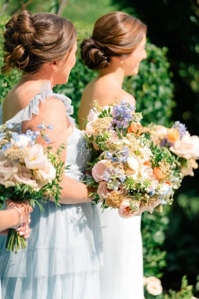 Bridesmaids with bouquets hold pastel wedding bouquets of roses, ranunculus, and blue delphinium with greenery in sunlit garden backdrop