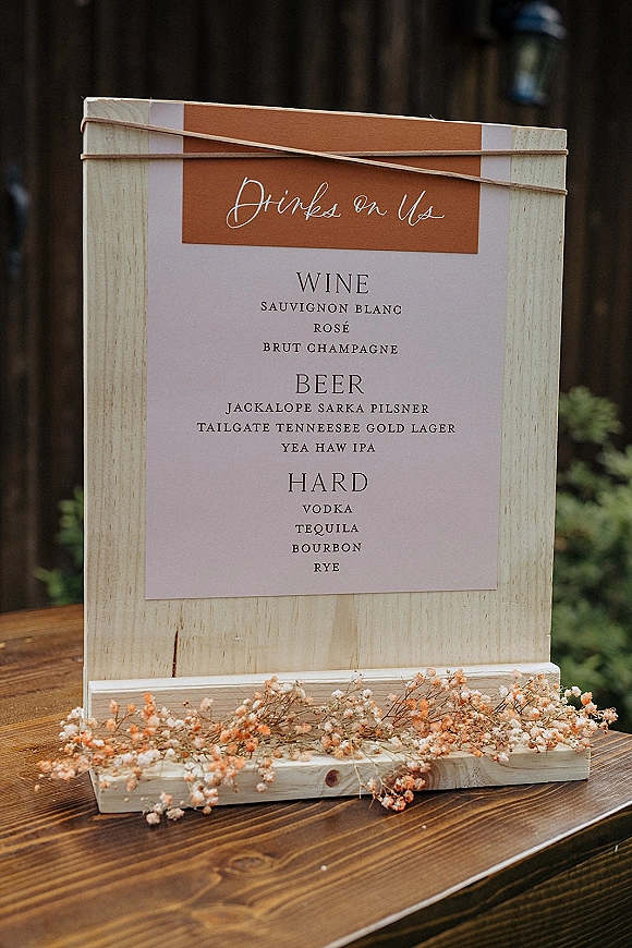 Wedding bar sign with a wedding drink menu sign on a wood stand, tied with string and dried flowers on a wood table outdoors