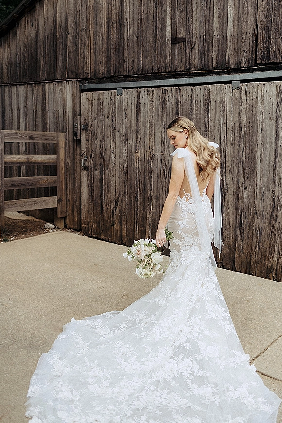 Bridal portrait of a bride in an open back wedding dress with lace and long train, holding a bouquet by a rustic wooden barn wall