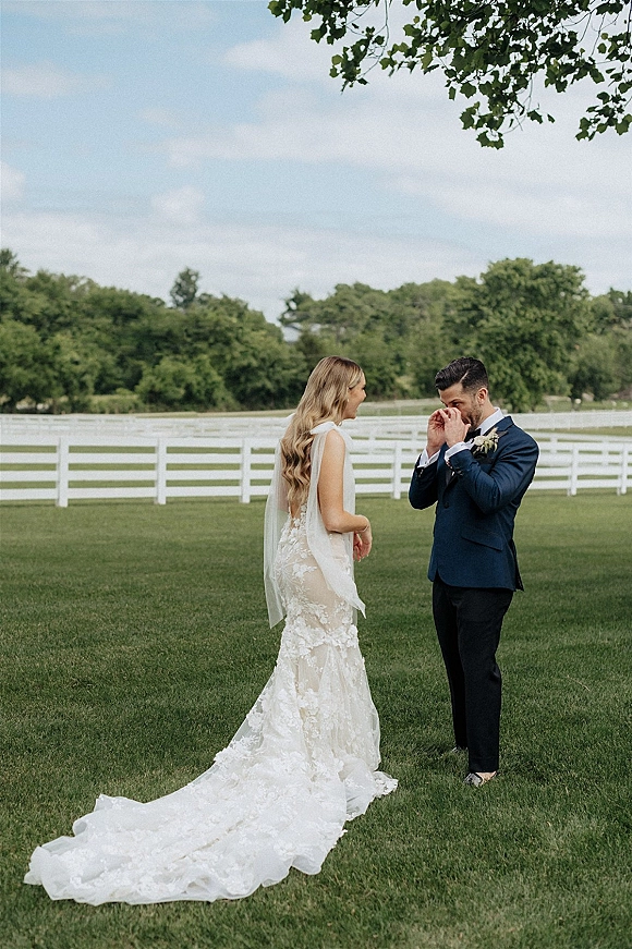 Wedding first look as bride in a lace back gown with long train and cape veil approaches groom on a green lawn by a white fence