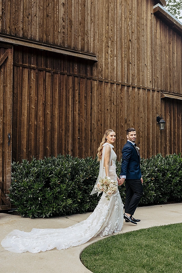 Couple portrait of bride and groom walking hand in hand, bride looking back as her lace train trails beside a rustic barn wall