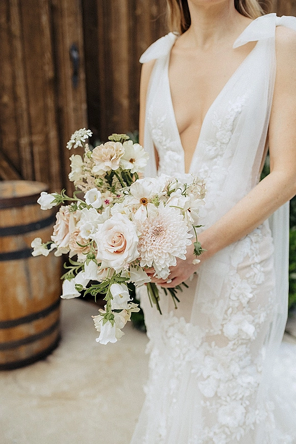 Bridal bouquet of white and blush flowers with garden roses, dahlias, and greenery held against a lace deep V gown by barn doors