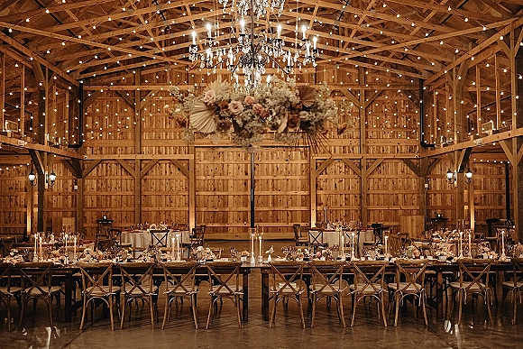 Barn reception decor with string lights and chandeliers over long tables, taper candles and neutral florals in a wood barn interior