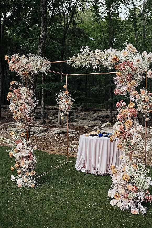 Wedding ceremony arch with roses and baby’s breath, hanging florals, and ribbon bows, set in a woodland clearing with vow table and flutes