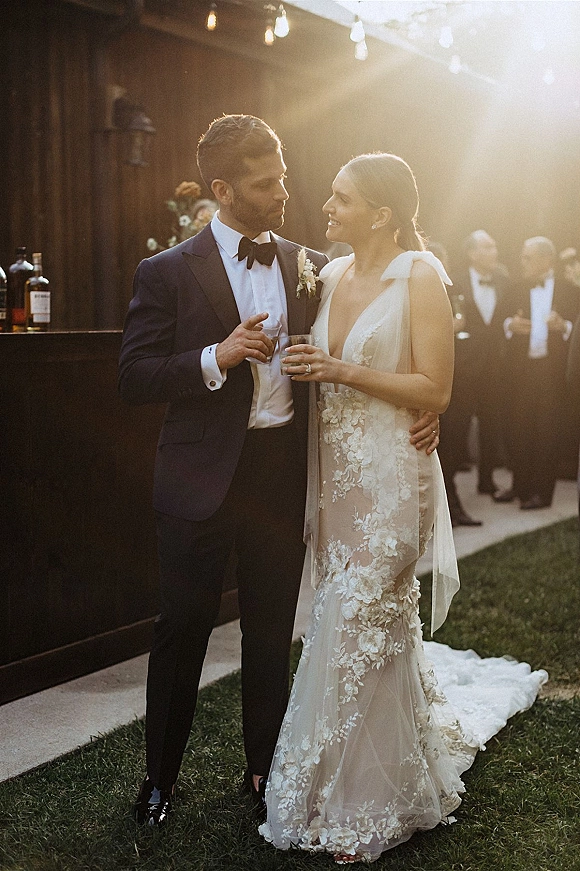 Couple portrait of bride and groom embracing, she holds a drink and he a whiskey glass at an outdoor bar under string lights