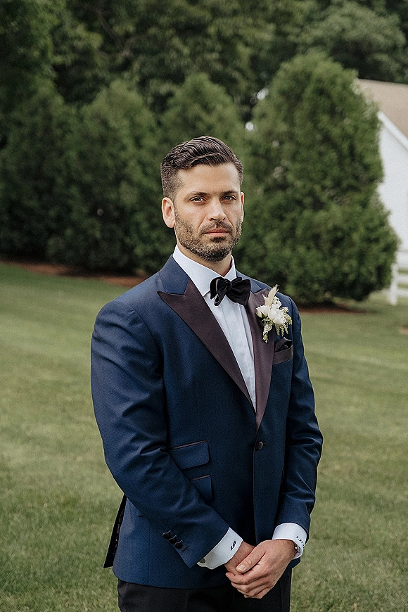 Groom portrait in a navy tuxedo with black bow tie, hands clasped, wearing a white rose boutonniere on a green lawn by a white fence