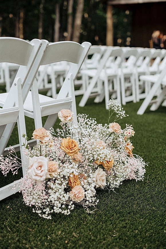 Ceremony aisle flowers in a ground arrangement of roses, carnations, and baby's breath beside white folding chairs on a grassy lawn with trees