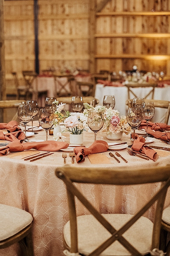 Reception tablescape with floral centerpieces, bud vases, rust napkins, and glowing votive candles on banquet tables in a wood barn interior