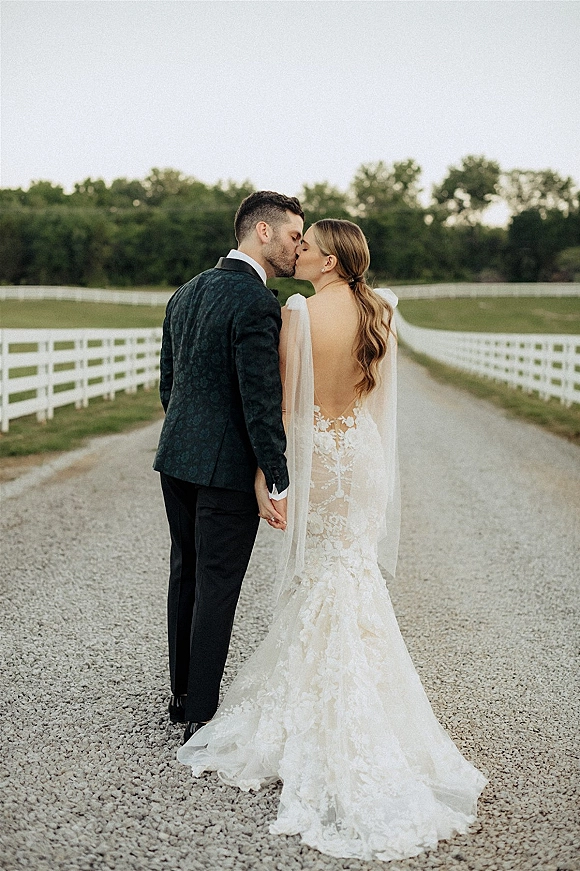 Wedding kiss portrait of bride and groom kissing, holding hands on a gravel road by a white fence, her cathedral veil flowing