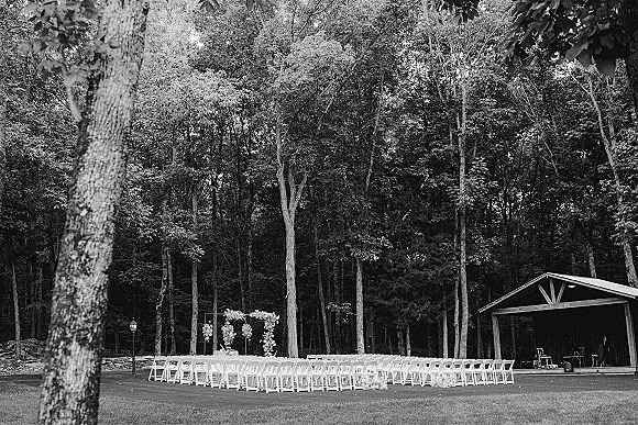 Ceremony setup for outdoor wedding ceremony with white folding chairs facing a floral altar under a pavilion beside wooded trees