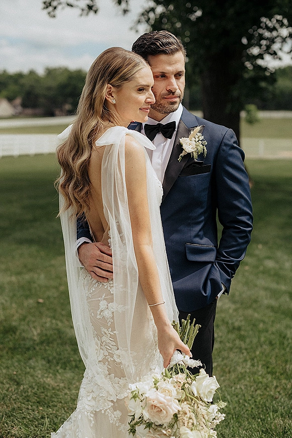 Couple portrait of bride and groom embrace in side profile, bride holding bouquet with veil and lace dress on a green lawn by a white fence