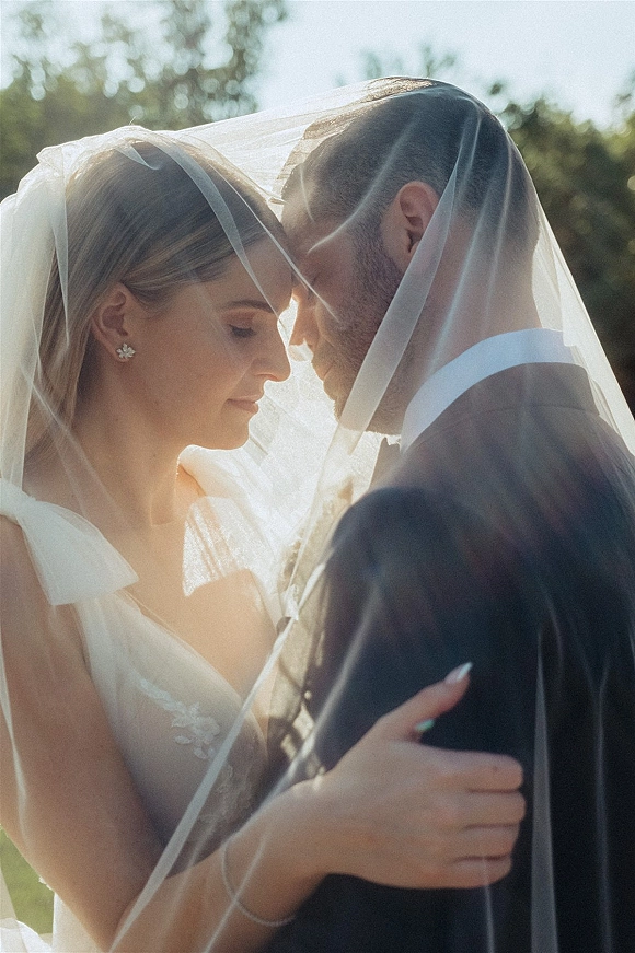 Wedding couple portrait with a veil over couple as bride and groom touch foreheads in an embrace amid outdoor greenery and sun flare