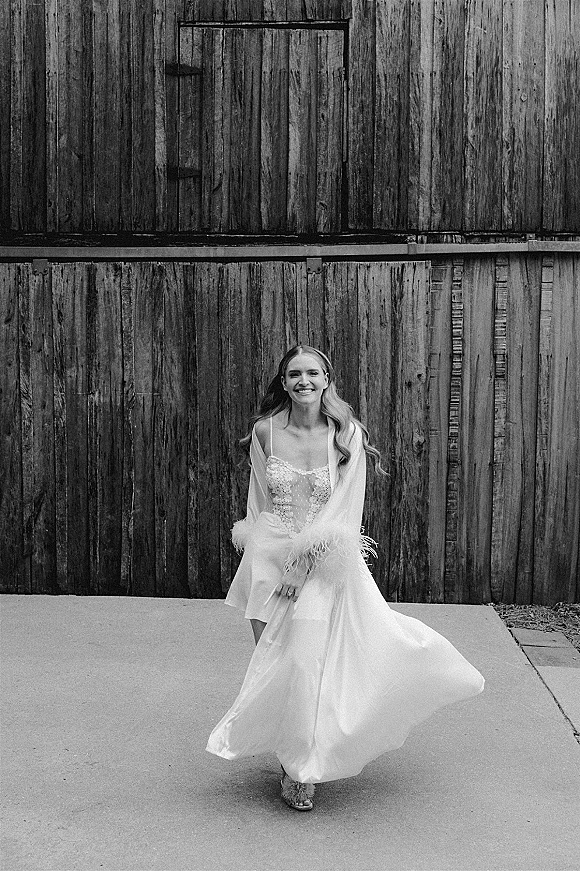 Bridal portrait of a smiling bride twirling her lace, spaghetti-strap wedding dress with feather-trim shawl against a rustic barn wall