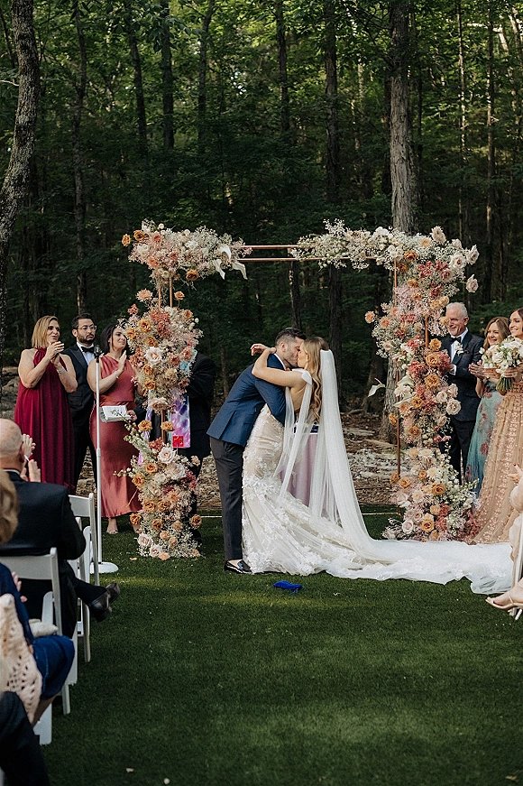 Ceremony kiss under a floral arch as the bride’s long veil drapes over her lace gown and guests applaud on a forest lawn outdoors