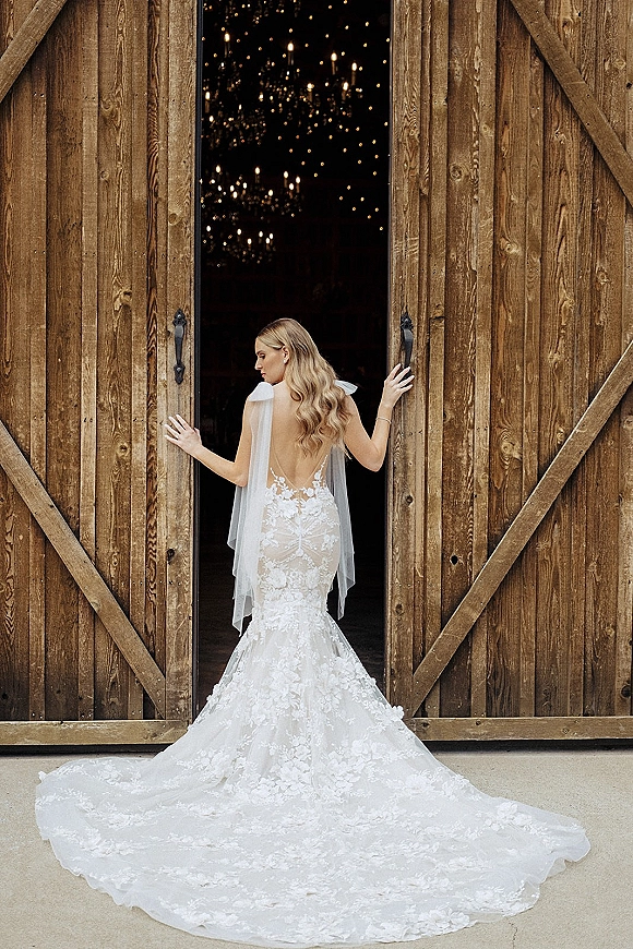 Bridal portrait of a bride in a backless lace wedding dress with cathedral veil and tulle train, standing at rustic barn doors under chandeliers