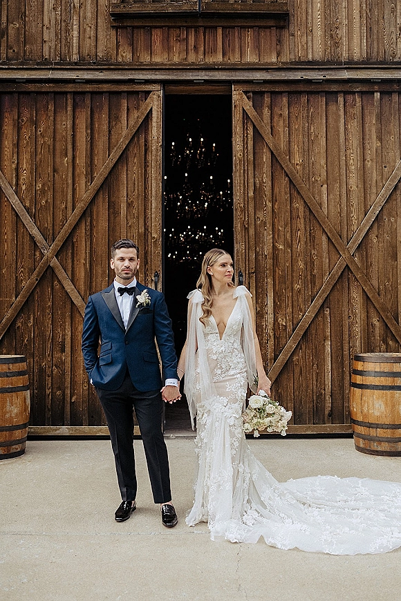 Couple portrait of bride and groom holding hands, her lace cathedral train and white rose bouquet by open barn doors with barrels