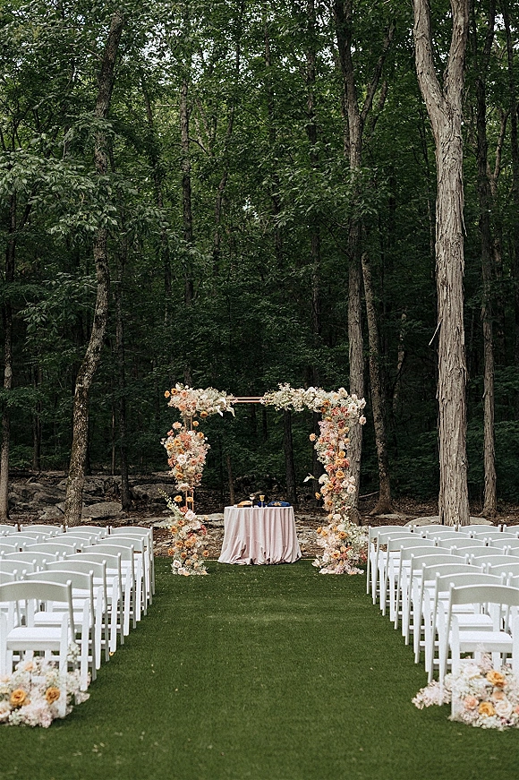 Ceremony setup for an outdoor wedding ceremony with white folding chairs lining an aisle to a pastel floral arch on a forest lawn