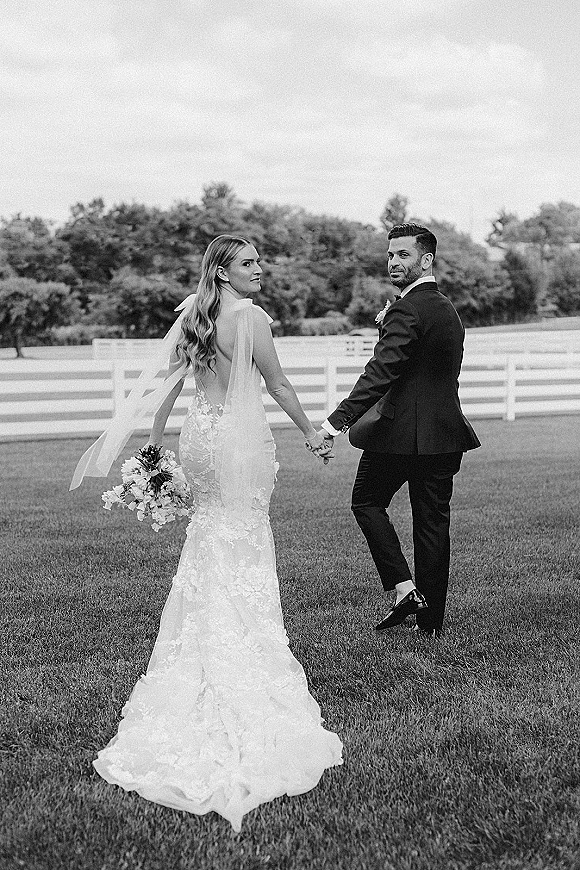 Couple portrait of bride and groom holding hands, walking away as she looks back, lace dress and long veil on a lawn by a white fence