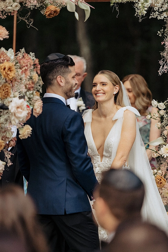 Ceremony moment as bride and groom at altar hold hands under a rose-and-greenery floral arch, with veil and guests by tree-lined aisle
