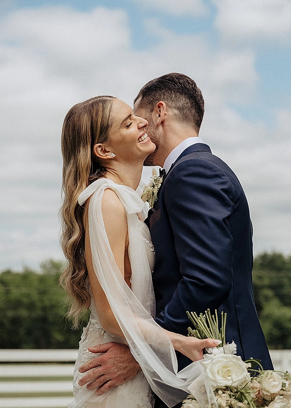 Wedding couple portrait with bride laughing as groom kisses her cheek, holding a white rose bouquet under blue sky with clouds and trees