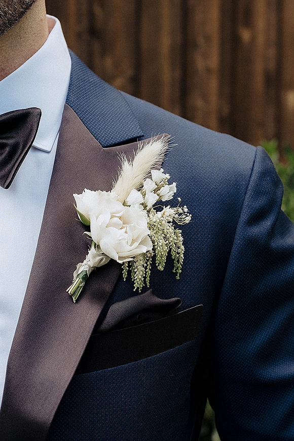 Groom boutonniere with a white rose boutonniere, greenery and pampas grass pinned to a tuxedo lapel, wood fence behind
