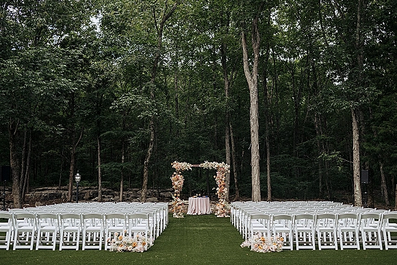 Ceremony setup for an outdoor wedding ceremony with white folding chairs lining a center aisle toward a floral arch on a woodland lawn