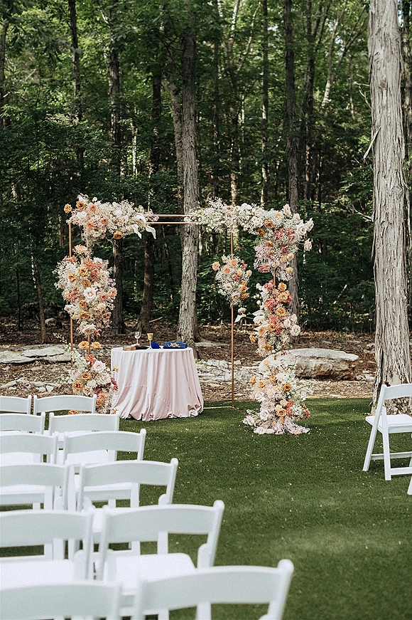 Ceremony setup with a copper floral arch, pastel garden roses, white chairs, and a blush table holding vows and champagne in woodland clearing