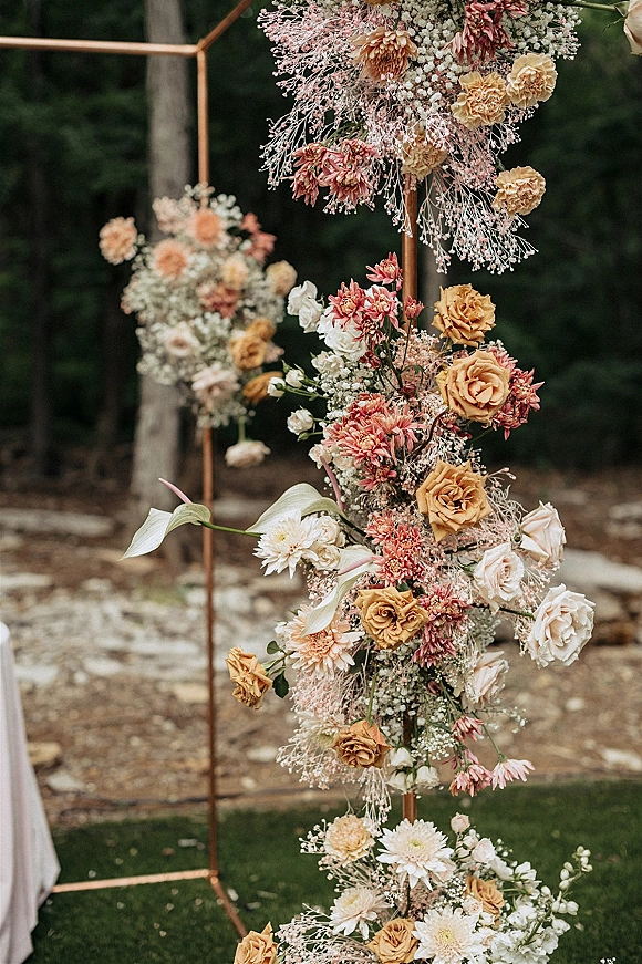 Wedding arch flowers on a copper geometric arch with asymmetrical roses, chrysanthemums, baby's breath, calla lilies, and greenery outdoors