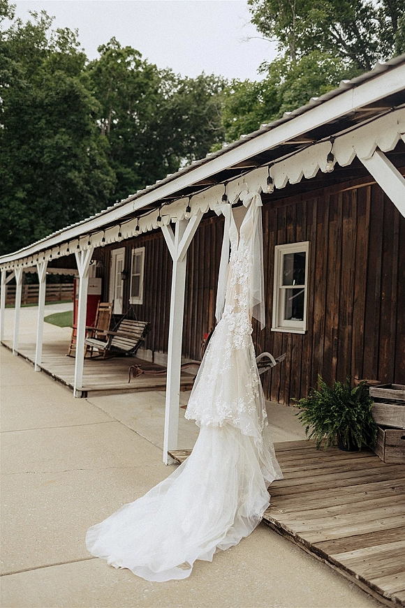 Wedding dress, lace wedding dress hanging from a porch post with long train, string lights, rocking chair, and potted fern on rustic barn porch