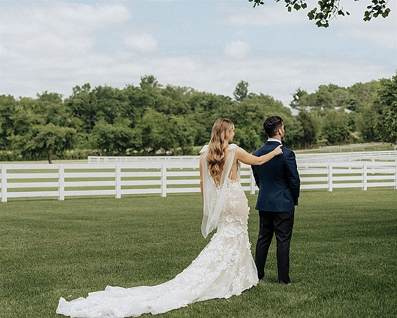 First look moment as bride taps groom’s shoulder, her lace open-back gown and veil trailing on a lawn by a white fence