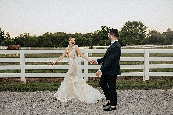 Couple portrait of bride and groom holding hands, her veil blowing in wind by a white fence with horses in a pasture backdrop