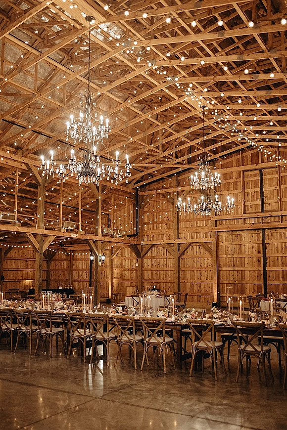 Reception decor in a barn wedding reception with long farmhouse tables, taper candles and floral centerpieces under string lights and chandeliers.