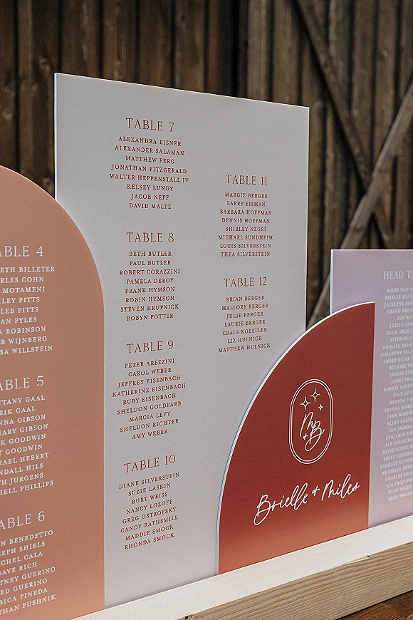 Wedding seating chart with wedding escort cards in arched printed guest list cards on a stand, featuring monogram and script names on a barn wood wall
