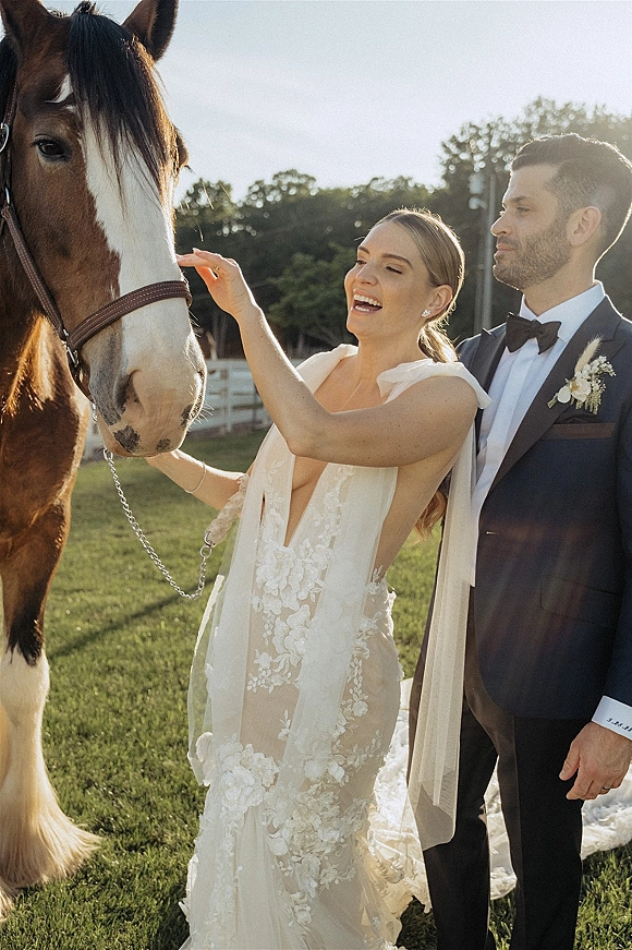 Couple portrait with bride petting horse, groom in tuxedo beside her as she holds the lead rope on a sunlit lawn by a fence